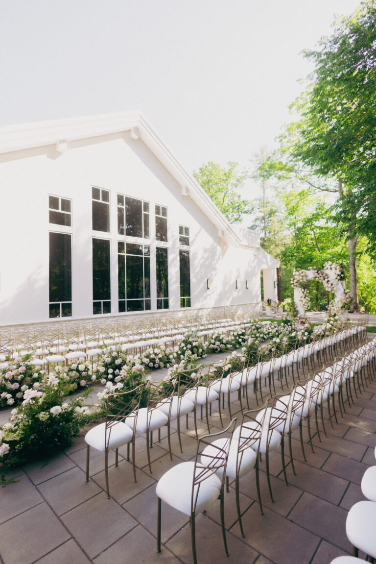 Outdoor wedding ceremony at Lakeview Pavilion in Foxborough, Massachusetts featuring white ceremony chairs, elegant floral aisle arrangements, and modern venue architecture surrounded by lush greenery.
