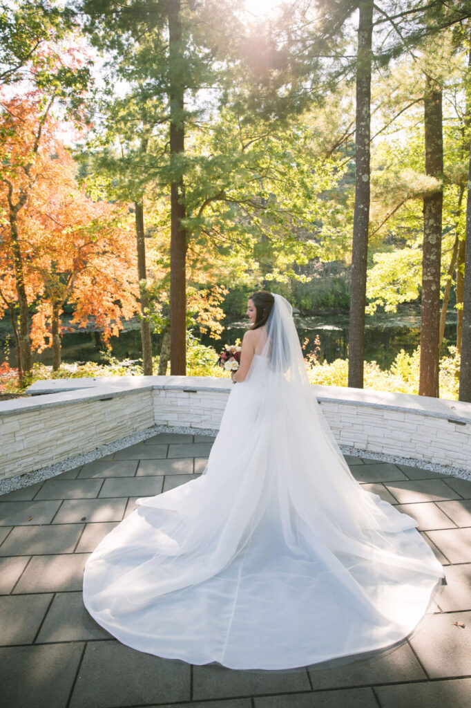 Bride in a flowing cathedral veil overlooking the waterfront at Lakeview Pavilion in Foxborough, Massachusetts, surrounded by vibrant fall foliage and scenic woodland views.