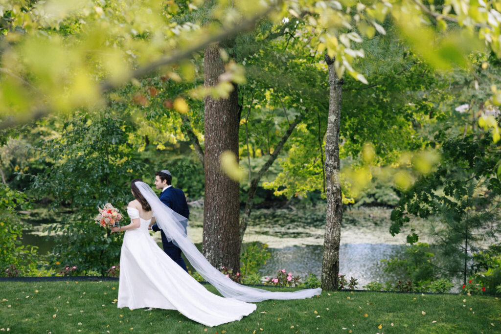 Bride and groom walking beside the waterfront at Lakeview Pavilion surrounded by lush trees and scenic lake views in Foxborough, Massachusetts.