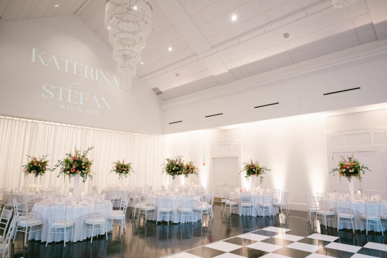 Elegant luxury wedding reception in the Lakeside Ballroom at Lakeview Pavilion featuring soaring ceilings, romantic white drapery, crystal chandeliers, soft uplighting, tall floral centerpieces, a black and white dance floor, and a personalized monogram projection in Foxborough, Massachusetts.