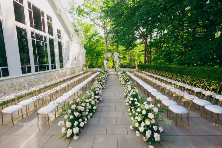 Outdoor wedding ceremony at Lakeview Pavilion featuring a floral aisle, elegant white rose arch, and gold guest chairs surrounded by lush trees in Foxborough, Massachusetts.